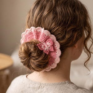 May include: A circular crochet hair accessory featuring a ruffled edge in light pink and a darker pink center. A small, three-dimensional crochet butterfly in shades of pink is attached to the side of the accessory, which is worn around a hair bun.