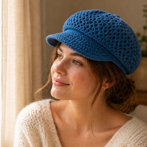 Peut inclure: Casquette gavroche bleue en crochet avec une petite visière. Le chapeau présente un motif ajouré. La casquette est portée par une personne portant un pull crème. L'arrière-plan comprend une fenêtre et un vase avec de la verdure.