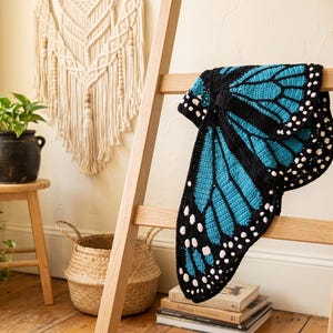 K&ouml;nnte beinhalten: Eine H&auml;keldecke, die wie Schmetterlingsfl&uuml;gel gestaltet ist und ein leuchtend blaues und schwarzes Farbschema mit wei&szlig;en Punkten an den R&auml;ndern aufweist. Das Textil ist &uuml;ber eine Holzleiter drapiert und zeigt die komplexe Garnarbeit.