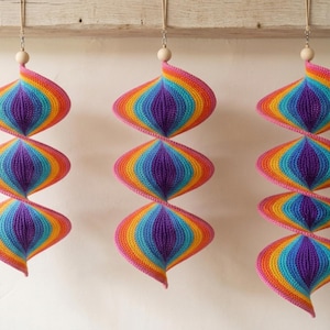 May include: Three hanging spiral ornaments crafted from crocheted yarn in a vibrant rainbow gradient of purple, blue, green, yellow, orange, and pink. Each piece features a natural wooden bead at the top and is suspended by a thin beige cord.