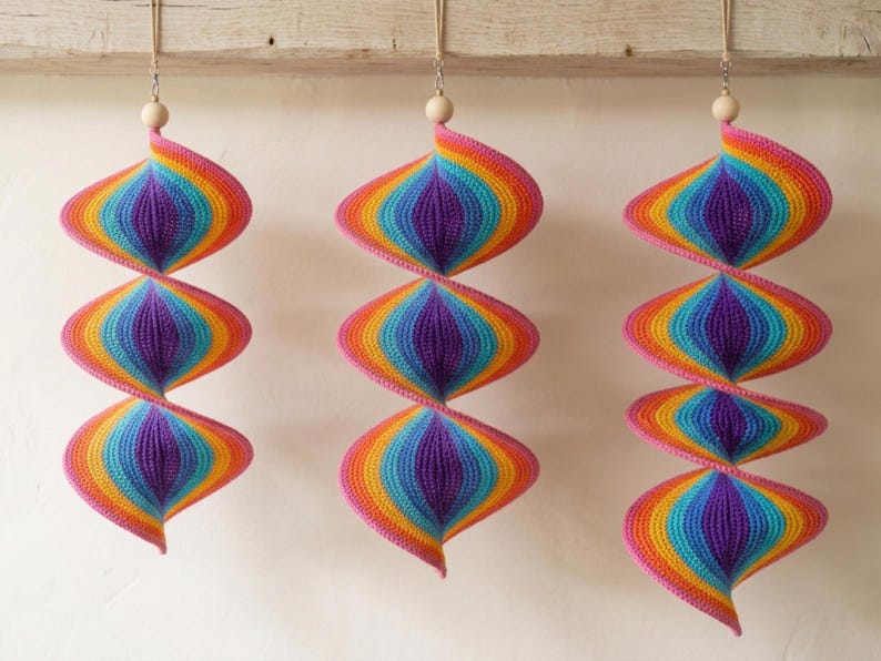 May include: Three hanging spiral ornaments crafted from crocheted yarn in a vibrant rainbow gradient of purple, blue, green, yellow, orange, and pink. Each piece features a natural wooden bead at the top and is suspended by a thin beige cord.