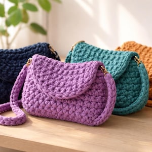 Puede incluir: Una colección de cuatro bolsos bandolera de ganchillo en colores sólidos que incluyen azul marino, lavanda, verde azulado y amarillo mostaza. Cada bolso presenta un patrón de tejido grueso y texturizado, una solapa redondeada y una correa trenzada.
