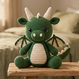 May include: A crocheted dragon plush toy featuring a dark green body with a cream-colored belly, wings, horns, and tail spikes. The creature has large black eyes, a rounded snout with two small cream nostrils, and sits in an upright position.