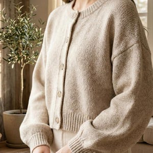 Op de afbeelding: Een beige vest met knopen, een ronde hals en geribde manchetten en zoom. De trui heeft lange mouwen en een iets kortere pasvorm. De stof lijkt een zachte brei te zijn. De achtergrond omvat een plant in een pot en een rieten mand.