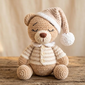 Könnte beinhalten: Ein gehäkelter Teddybär aus hellbraunem Garn, der eine passende Schlafmütze mit weißem Bommel und weißem Rand trägt. Der Bär hat einen weißen Kragen, drei kleine Knöpfe auf der Brust und geschlossene Augen, die mit schwarzem Faden gestickt sind.