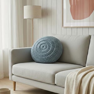 Puede incluir: Un coj&iacute;n redondo azul con un dise&ntilde;o en espiral descansa sobre un sof&aacute; gris claro. Una manta beige est&aacute; sobre el sof&aacute;. Una l&aacute;mpara de pie y una obra de arte est&aacute;n en el fondo, creando un ambiente acogedor en la sala de estar.