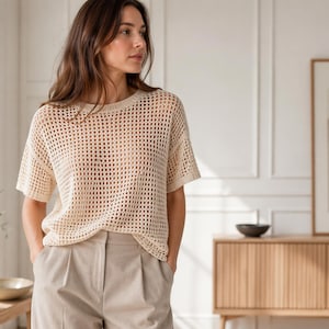 Pode incluir: Um top de malha creme com tecido aberto, mangas curtas e gola redonda. O top é combinado com calças plissadas bege claro. O conjunto é casual e confortável.