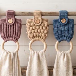 May include: Three hanging kitchen towel holders featuring crocheted tops in mauve, beige, and blue, each secured with a wooden button. Each holder includes a smooth, circular wooden ring that supports a white fabric towel hanging vertically.