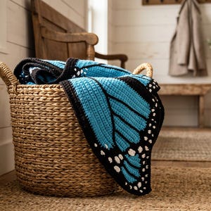 K&ouml;nnte beinhalten: Ein geflochtener Seegraskorb mit einer geh&auml;kelten Decke, die ein leuchtend blau-schwarzes Schmetterlingsfl&uuml;gelmuster mit wei&szlig;en Akzenten an den R&auml;ndern aufweist. Die Decke ist gefaltet und &uuml;ber den Rand des runden, strukturierten Aufbewahrungskorbs drapiert.