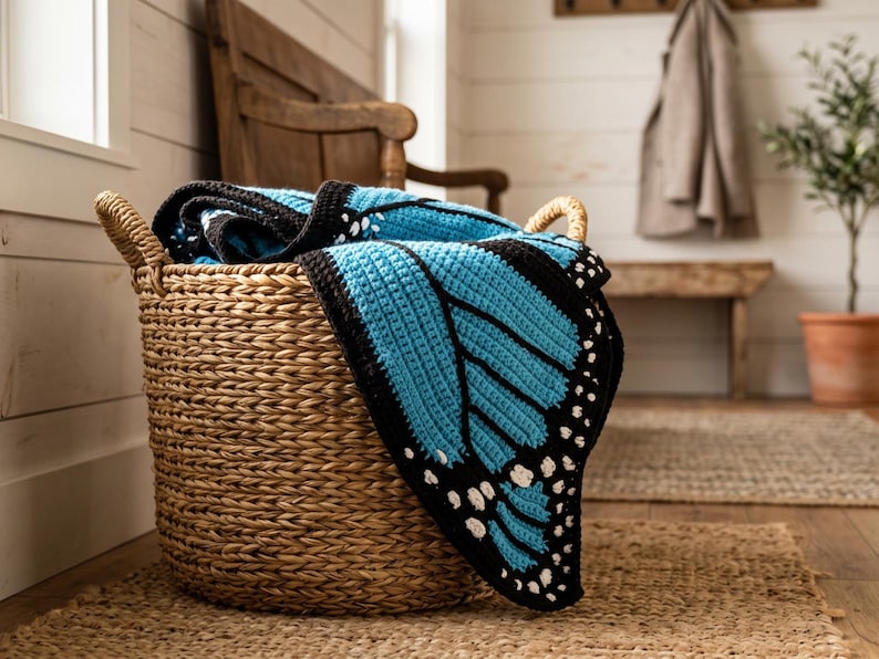 K&ouml;nnte beinhalten: Ein geflochtener Seegraskorb mit einer geh&auml;kelten Decke, die ein leuchtend blau-schwarzes Schmetterlingsfl&uuml;gelmuster mit wei&szlig;en Akzenten an den R&auml;ndern aufweist. Die Decke ist gefaltet und &uuml;ber den Rand des runden, strukturierten Aufbewahrungskorbs drapiert.