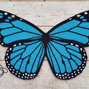 K&ouml;nnte beinhalten: Ein gro&szlig;es, flaches, geh&auml;keltes Textil in Form von Schmetterlingsfl&uuml;geln mit leuchtend blauen Abschnitten, die schwarz umrandet sind und kleine wei&szlig;e Punkte an den R&auml;ndern aufweisen. Eine Holzh&auml;kelnadel und ein kleines Kn&auml;uel schwarzes Garn liegen links.