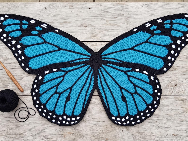 K&ouml;nnte beinhalten: Ein gro&szlig;es, flaches, geh&auml;keltes Textil in Form von Schmetterlingsfl&uuml;geln mit leuchtend blauen Abschnitten, die schwarz umrandet sind und kleine wei&szlig;e Punkte an den R&auml;ndern aufweisen. Eine Holzh&auml;kelnadel und ein kleines Kn&auml;uel schwarzes Garn liegen links.