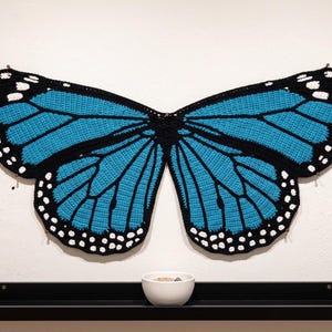 K&ouml;nnte beinhalten: Ein gro&szlig;es, flaches Wandbehang-Objekt aus geh&auml;kelten Schmetterlingsfl&uuml;geln mit einem leuchtend blauen und schwarzen Muster sowie wei&szlig;en Punkten an den R&auml;ndern. Das Textilst&uuml;ck ist an die Wand gepinnt, &uuml;ber einem schwarzen Regal mit einer wei&szlig;en Schale.