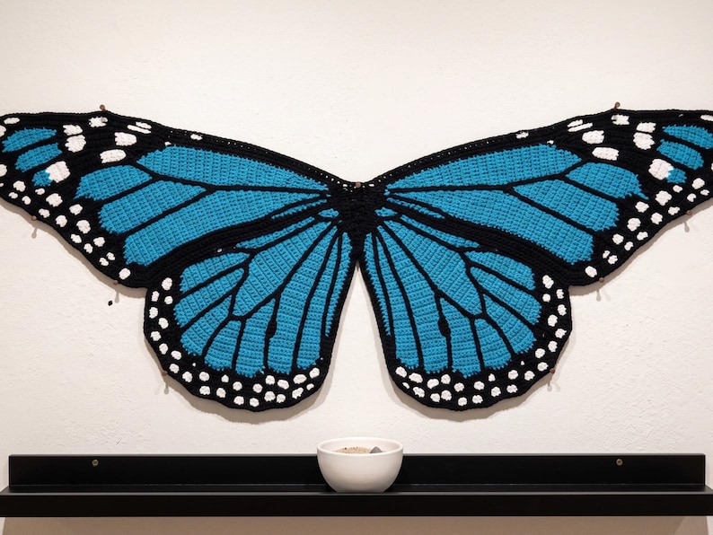 K&ouml;nnte beinhalten: Ein gro&szlig;es, flaches Wandbehang-Objekt aus geh&auml;kelten Schmetterlingsfl&uuml;geln mit einem leuchtend blauen und schwarzen Muster sowie wei&szlig;en Punkten an den R&auml;ndern. Das Textilst&uuml;ck ist an die Wand gepinnt, &uuml;ber einem schwarzen Regal mit einer wei&szlig;en Schale.