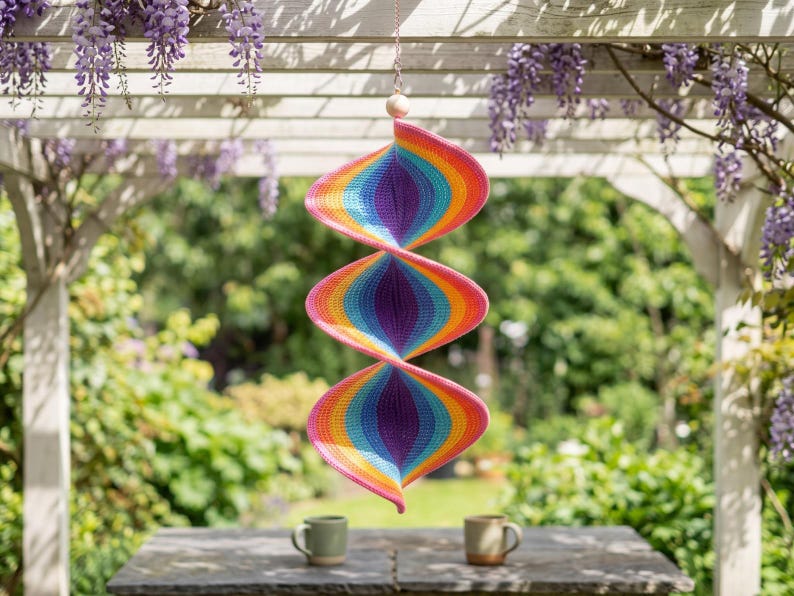 May include: A hanging spiral wind spinner featuring a vibrant crochet pattern with concentric rings of pink, orange, yellow, light blue, and purple. The structure consists of three connected, twisted sections suspended from a thin chain with a wooden bead.