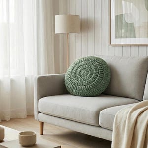Puede incluir: Un coj&iacute;n redondo de ganchillo verde salvia descansa sobre un sof&aacute; gris claro. El coj&iacute;n tiene un dise&ntilde;o en espiral. Una manta beige est&aacute; sobre el sof&aacute;. Una l&aacute;mpara de pie y una obra de arte est&aacute;n en el fondo. Decoraci&oacute;n del hogar.