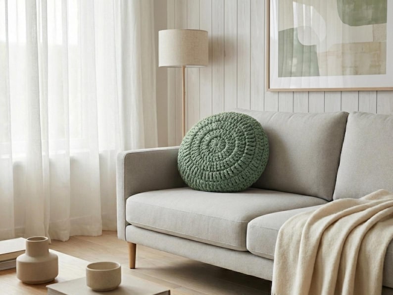 Puede incluir: Un coj&iacute;n redondo de ganchillo verde salvia descansa sobre un sof&aacute; gris claro. El coj&iacute;n tiene un dise&ntilde;o en espiral. Una manta beige est&aacute; sobre el sof&aacute;. Una l&aacute;mpara de pie y una obra de arte est&aacute;n en el fondo. Decoraci&oacute;n del hogar.