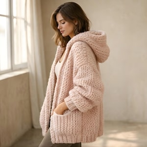 Puede incluir: Un cárdigan de punto grueso de color rosa claro con capucha y bolsillos delanteros. El suéter de gran tamaño está hecho de un hilo grueso y texturizado. La modelo lleva un top blanco debajo del cárdigan.