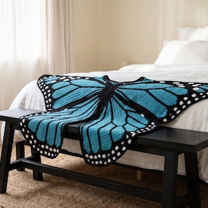 K&ouml;nnte beinhalten: Eine geh&auml;kelte Decke in Schmetterlingsform mit leuchtend blauen Fl&uuml;geln, schwarzen R&auml;ndern und wei&szlig;en Punkten an den Kanten. Das Textil zeigt ein detailliertes Fl&uuml;gelmuster mit schwarzen Adern und einem Mittelteil, das zum &Uuml;berlegen gedacht ist.