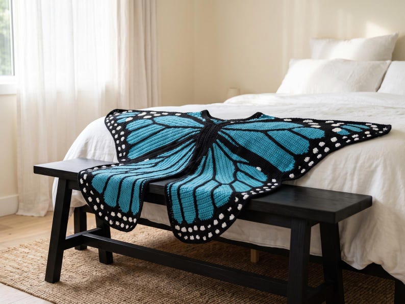 K&ouml;nnte beinhalten: Eine geh&auml;kelte Decke in Schmetterlingsform mit leuchtend blauen Fl&uuml;geln, schwarzen R&auml;ndern und wei&szlig;en Punkten an den Kanten. Das Textil zeigt ein detailliertes Fl&uuml;gelmuster mit schwarzen Adern und einem Mittelteil, das zum &Uuml;berlegen gedacht ist.
