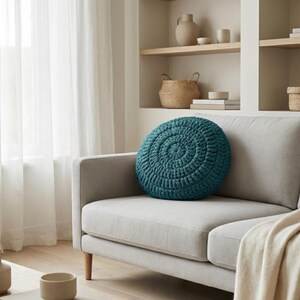 Puede incluir: Un coj&iacute;n redondo de ganchillo color turquesa descansa sobre un sof&aacute; gris claro. El coj&iacute;n tiene un dise&ntilde;o en espiral. El sof&aacute; est&aacute; en una habitaci&oacute;n bien iluminada con una paleta de colores neutros, con una manta beige y patas de madera.