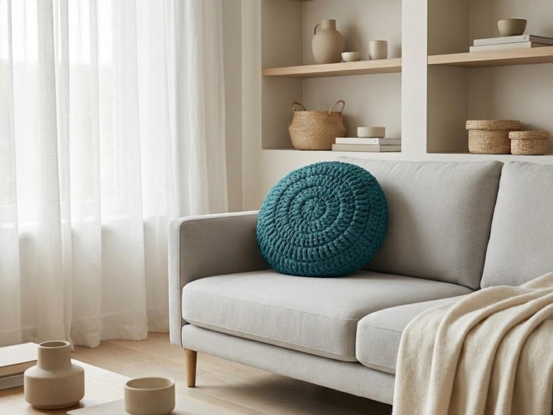 Puede incluir: Un coj&iacute;n redondo de ganchillo color turquesa descansa sobre un sof&aacute; gris claro. El coj&iacute;n tiene un dise&ntilde;o en espiral. El sof&aacute; est&aacute; en una habitaci&oacute;n bien iluminada con una paleta de colores neutros, con una manta beige y patas de madera.