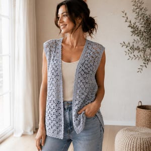 May include: A light blue, sleeveless, open-front crochet vest. The vest has a delicate, openwork pattern and is worn over a white tank top and blue jeans. The vest is a stylish layering piece.