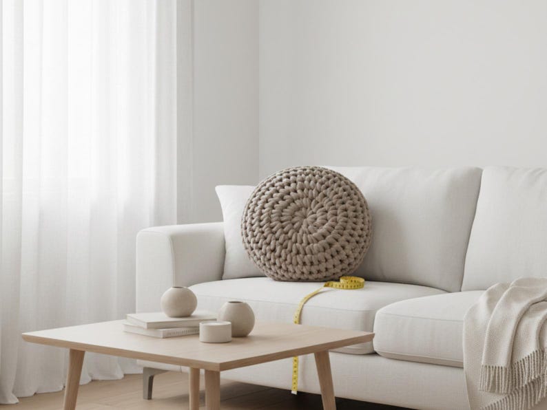 Puede incluir: Un sal&oacute;n de tonos neutros con un sof&aacute; blanco y un gran coj&iacute;n redondo beige tejido. Una mesa de centro de madera con jarrones decorativos y libros. Una cinta m&eacute;trica amarilla descansa sobre el sof&aacute;.