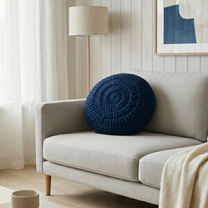 Puede incluir: Un coj&iacute;n redondo de ganchillo azul marino con un dise&ntilde;o en espiral descansa sobre un sof&aacute; gris claro. El coj&iacute;n a&ntilde;ade un toque de color a la sala de estar de tonos neutros. Una manta beige est&aacute; sobre el sof&aacute;.
