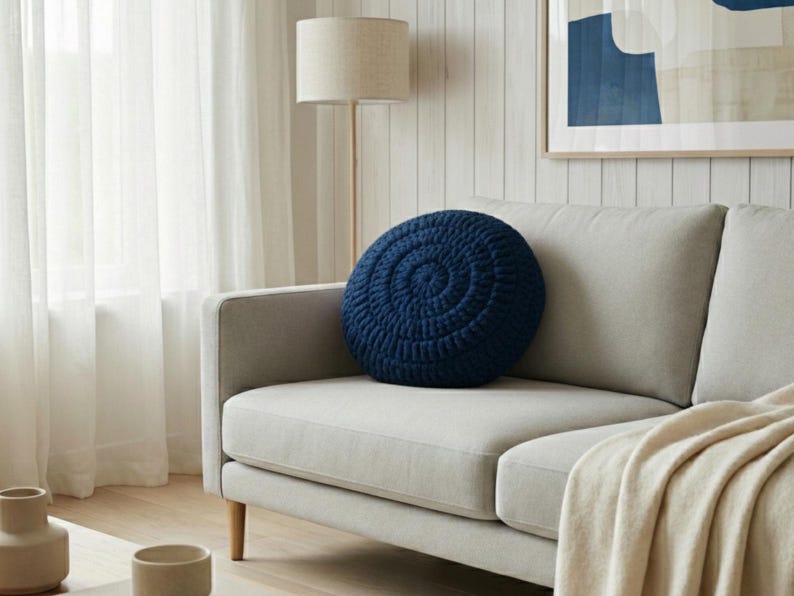 Puede incluir: Un coj&iacute;n redondo de ganchillo azul marino con un dise&ntilde;o en espiral descansa sobre un sof&aacute; gris claro. El coj&iacute;n a&ntilde;ade un toque de color a la sala de estar de tonos neutros. Una manta beige est&aacute; sobre el sof&aacute;.