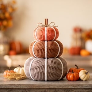 May include: A stacked set of three crocheted pumpkins in orange, brown, and taupe, each featuring white vertical yarn accents. The top pumpkin is topped with two cinnamon sticks and a tied twine bow, while smaller pumpkins sit at the base.