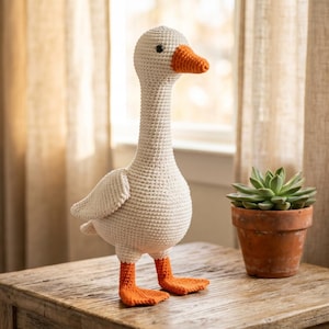 Op de afbeelding: Een handgemaakt gehaakt gansspeeltje met een crèmekleurig lichaam, een lange nek en een gestructureerd steekpatroon. De gans heeft een feloranje snavel en bijpassende oranje zwemvliezen, met kleine zwarte kraaloogjes aan de zijkanten van de kop.
