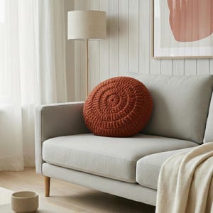 Puede incluir: Un coj&iacute;n redondo de ganchillo color &oacute;xido con un dise&ntilde;o en espiral descansa sobre un sof&aacute; gris claro. El sof&aacute; tiene patas de madera y una manta color crema. Una l&aacute;mpara de pie y una obra de arte est&aacute;n en el fondo.