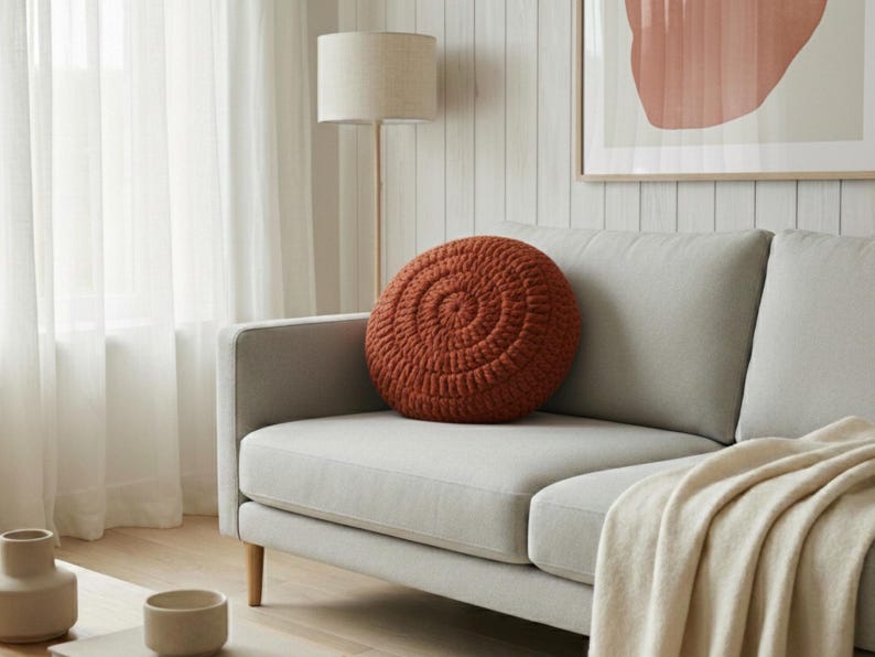 Puede incluir: Un coj&iacute;n redondo de ganchillo color &oacute;xido con un dise&ntilde;o en espiral descansa sobre un sof&aacute; gris claro. El sof&aacute; tiene patas de madera y una manta color crema. Una l&aacute;mpara de pie y una obra de arte est&aacute;n en el fondo.