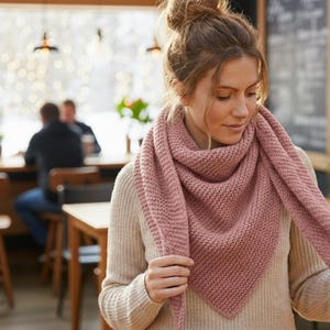Può includere: Una donna indossa una sciarpa lavorata a maglia color rosa antico. La sciarpa è triangolare e presenta un motivo strutturato. Indossa un maglione beige chiaro. Lo sfondo mostra un ambiente da caffè con tavoli e sedie.