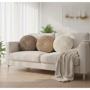 May include: A cream-colored sofa with three round, textured pillows in shades of brown and white. A matching throw blanket is draped over the arm. Potted plants and a framed abstract artwork complete the cozy living room scene.