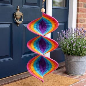 May include: A hanging spiral decoration made of crocheted yarn featuring a vibrant rainbow gradient pattern of pink, orange, yellow, green, blue, and purple. The structure consists of three connected, twisted sections suspended from a wooden bead.