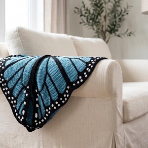 K&ouml;nnte beinhalten: Eine H&auml;keldecke im Design eines Schmetterlingsfl&uuml;gels mit einem leuchtend hellblauen Zentrum, schwarzen Adern und einem schwarzen Rand, der mit kleinen wei&szlig;en Punkten entlang der Au&szlig;enkante verziert ist, drapiert &uuml;ber einem cremefarbenen Sofa.