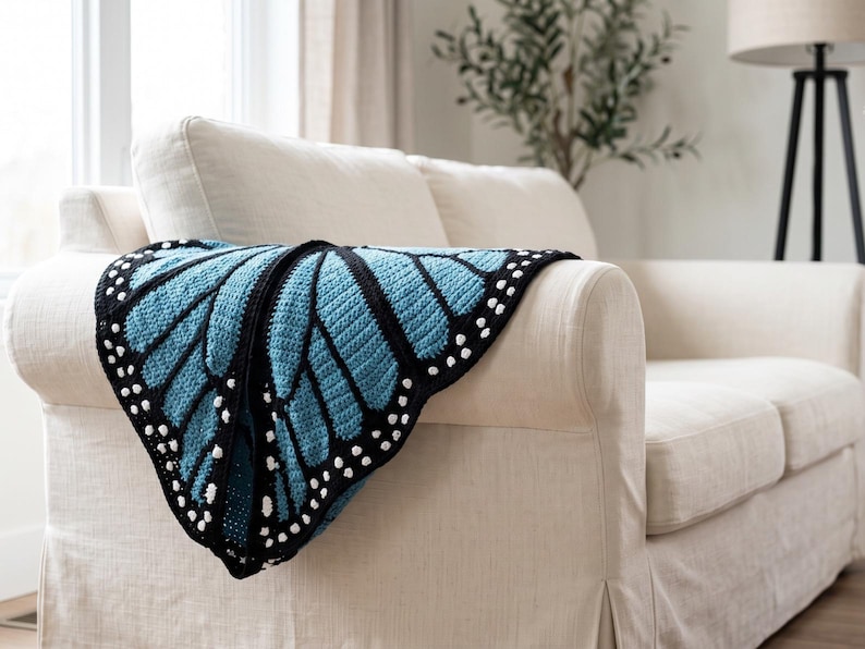 K&ouml;nnte beinhalten: Eine H&auml;keldecke im Design eines Schmetterlingsfl&uuml;gels mit einem leuchtend hellblauen Zentrum, schwarzen Adern und einem schwarzen Rand, der mit kleinen wei&szlig;en Punkten entlang der Au&szlig;enkante verziert ist, drapiert &uuml;ber einem cremefarbenen Sofa.