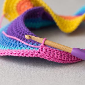 May include: A multi-colored crochet project featuring concentric rows of yellow, orange, purple, blue, and pink yarn. A wooden crochet hook with a purple handle is inserted into the pink edge of the work, holding a single loop of yarn in place.