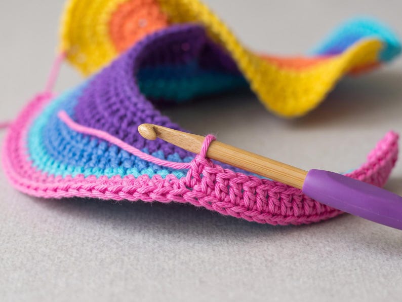 May include: A multi-colored crochet project featuring concentric rows of yellow, orange, purple, blue, and pink yarn. A wooden crochet hook with a purple handle is inserted into the pink edge of the work, holding a single loop of yarn in place.