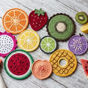 Puede incluir: Una colección de posavasos de ganchillo redondos y con formas que imitan varias frutas, incluyendo naranja, fresa, kiwi, pitahaya, limón, lima, sandía, melocotón, piña y rodajas de fruta morada, junto a trozos de fruta real.