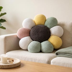 May include: A round, flower-shaped throw pillow featuring a dark brown, spiraled center surrounded by plush, textured petals in alternating colors of white, soft yellow, sage green, and dusty pink, crafted with a visible knit or crochet stitch.