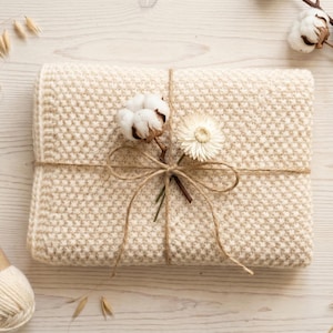 May include: A folded cream-colored knit blanket with a textured honeycomb pattern, tied with thin brown twine. A single white cotton boll and a small dried flower are tucked into the twine bow, accompanied by a ball of matching cream yarn.