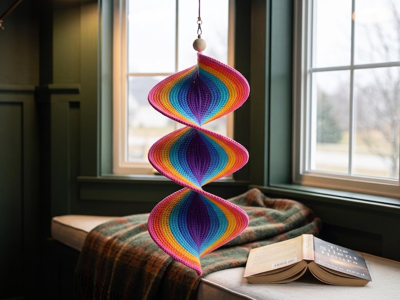 May include: A hanging spiral decoration featuring a vibrant crochet pattern with repeating bands of pink, orange, yellow, light blue, and dark purple. The structure consists of three connected, twisted sections suspended from a single white wooden bead.