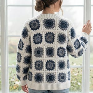 Può includere: Un maglione all'uncinetto color crema con un motivo quadrato di disegni floreali blu e grigi. Il maglione ha maniche lunghe e orlo e polsini a coste. La persona è in piedi davanti a una finestra.