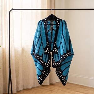 K&ouml;nnte beinhalten: Ein geh&auml;kelter Schal mit einem leuchtend blauen und schwarzen Schmetterlingsfl&uuml;gelmuster. Das Design umfasst schwarze R&auml;nder mit wei&szlig;en kreisf&ouml;rmigen Akzenten, die das Aussehen von Schmetterlingsfl&uuml;geln nachahmen, h&auml;ngend an einer schwarzen Kleiderstange.