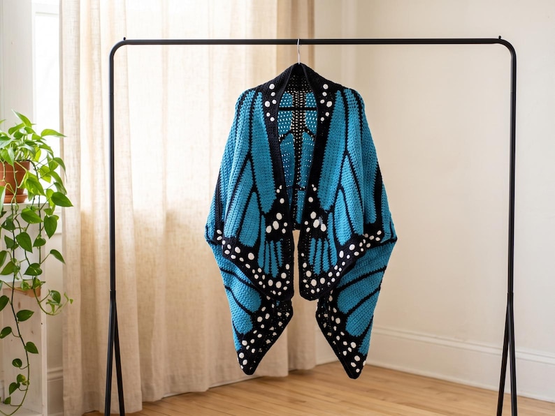 K&ouml;nnte beinhalten: Ein geh&auml;kelter Schal mit einem leuchtend blauen und schwarzen Schmetterlingsfl&uuml;gelmuster. Das Design umfasst schwarze R&auml;nder mit wei&szlig;en kreisf&ouml;rmigen Akzenten, die das Aussehen von Schmetterlingsfl&uuml;geln nachahmen, h&auml;ngend an einer schwarzen Kleiderstange.