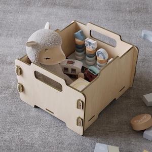 Può includere: Un contenitore per giocattoli in legno colorato chiaro pieno di vari giocattoli, tra cui una pecora di peluche, giocattoli impilabili in legno e veicoli in miniatura. La scatola ha maniglie ed è realizzata in legno colorato chiaro.