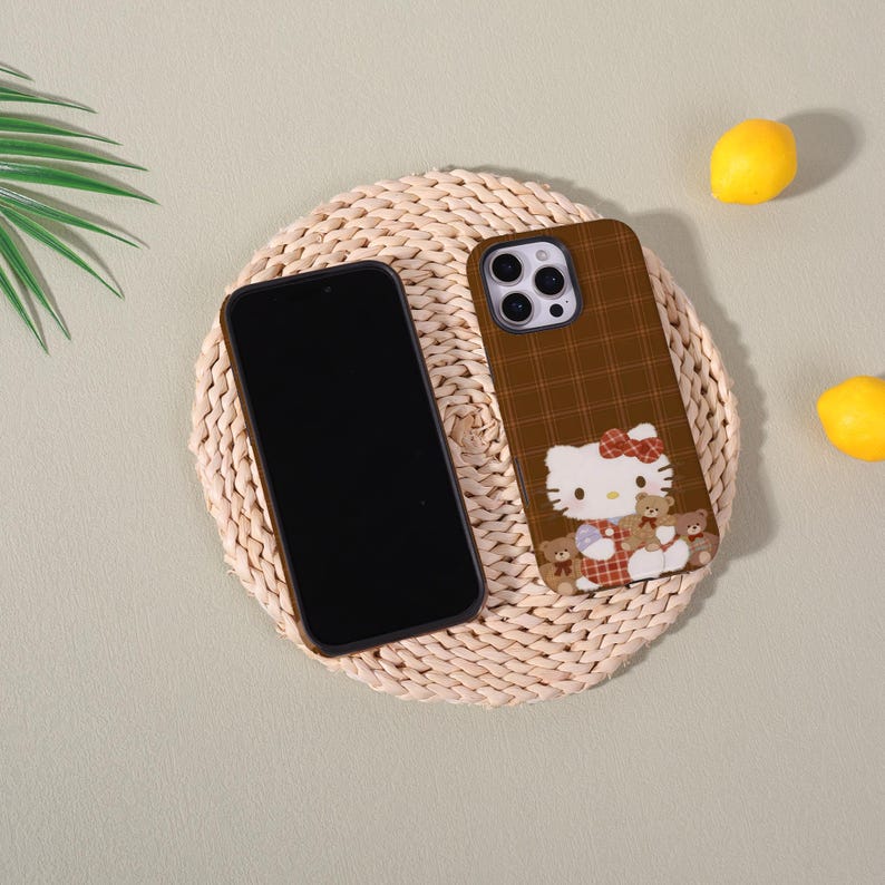 Op de afbeelding: Een bruine telefoonhoes met een Hello Kitty-ontwerp, met een rood ruitpatroon en teddyberen. De hoes ligt naast een zwarte smartphone op een geweven mat, met citroenen op de achtergrond.